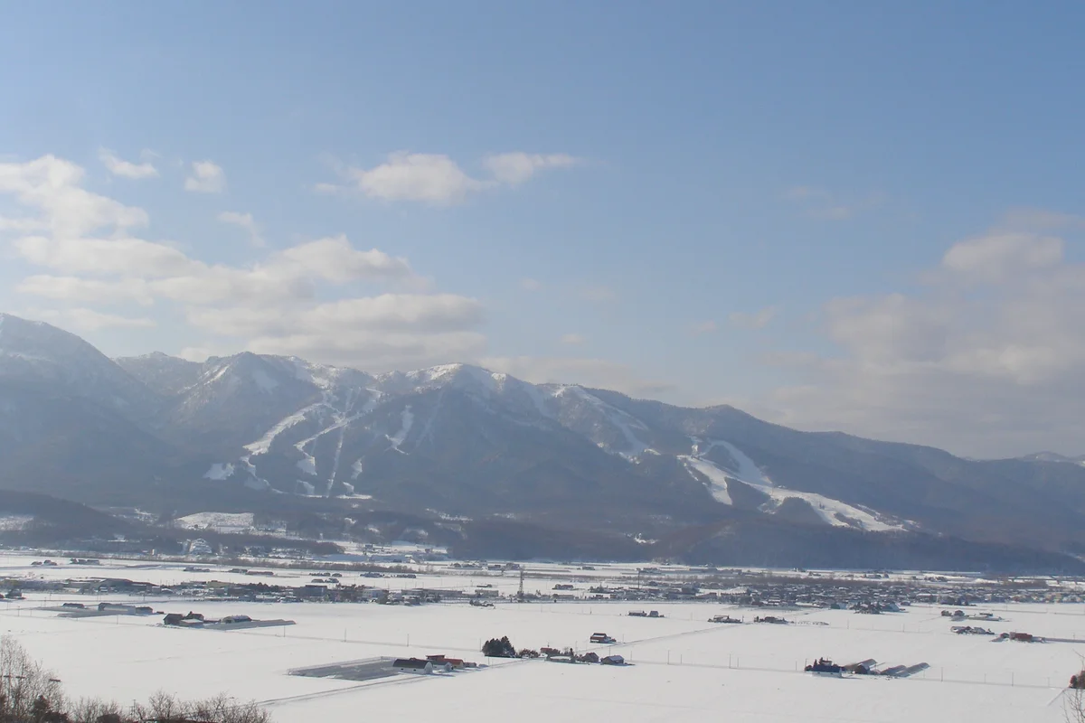 Campi innevati e la cittadina nella valle; dietro, montagne coperte di bosco solcate dalle piste bianche della stazione sciistica di Furano, sotto un cielo azzurro