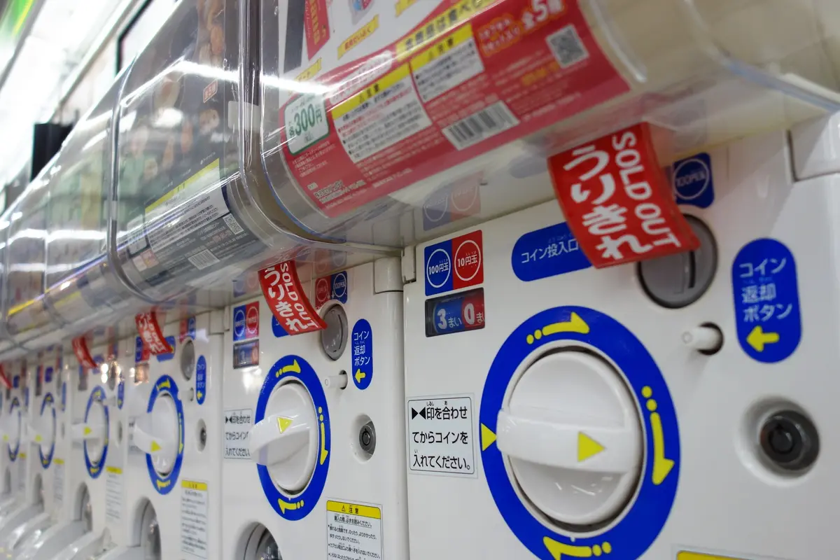 Una fila di macchine per le capsule giapponesi, ognuna con fessura per le monete, tasto di restituzione e una grande manopola bianca