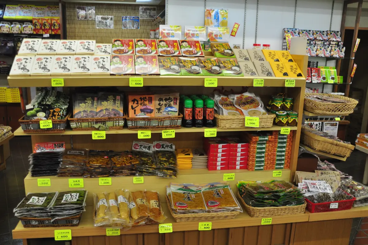 An omiyage food counter in Beppu stacked with boxed dried shiitake, local curry and pickles, each under a small yellow price tag