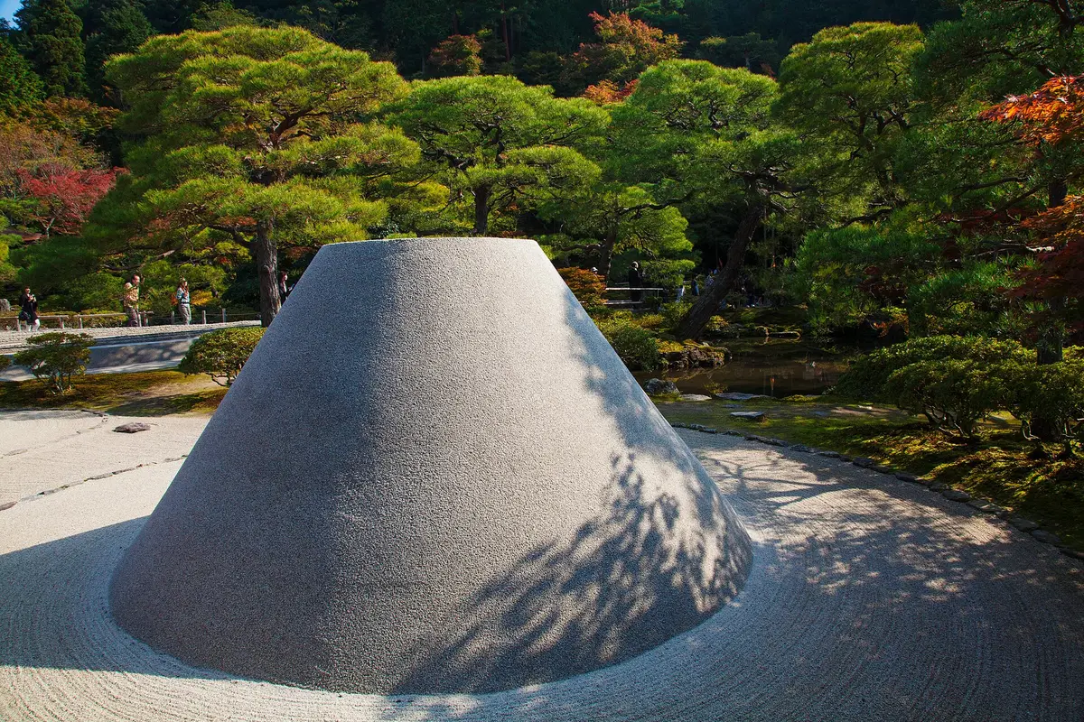 A flat-topped cone of pale sand, the Kogetsudai, standing in a bed of raked sand at Ginkaku-ji, with pines and autumn maples behind