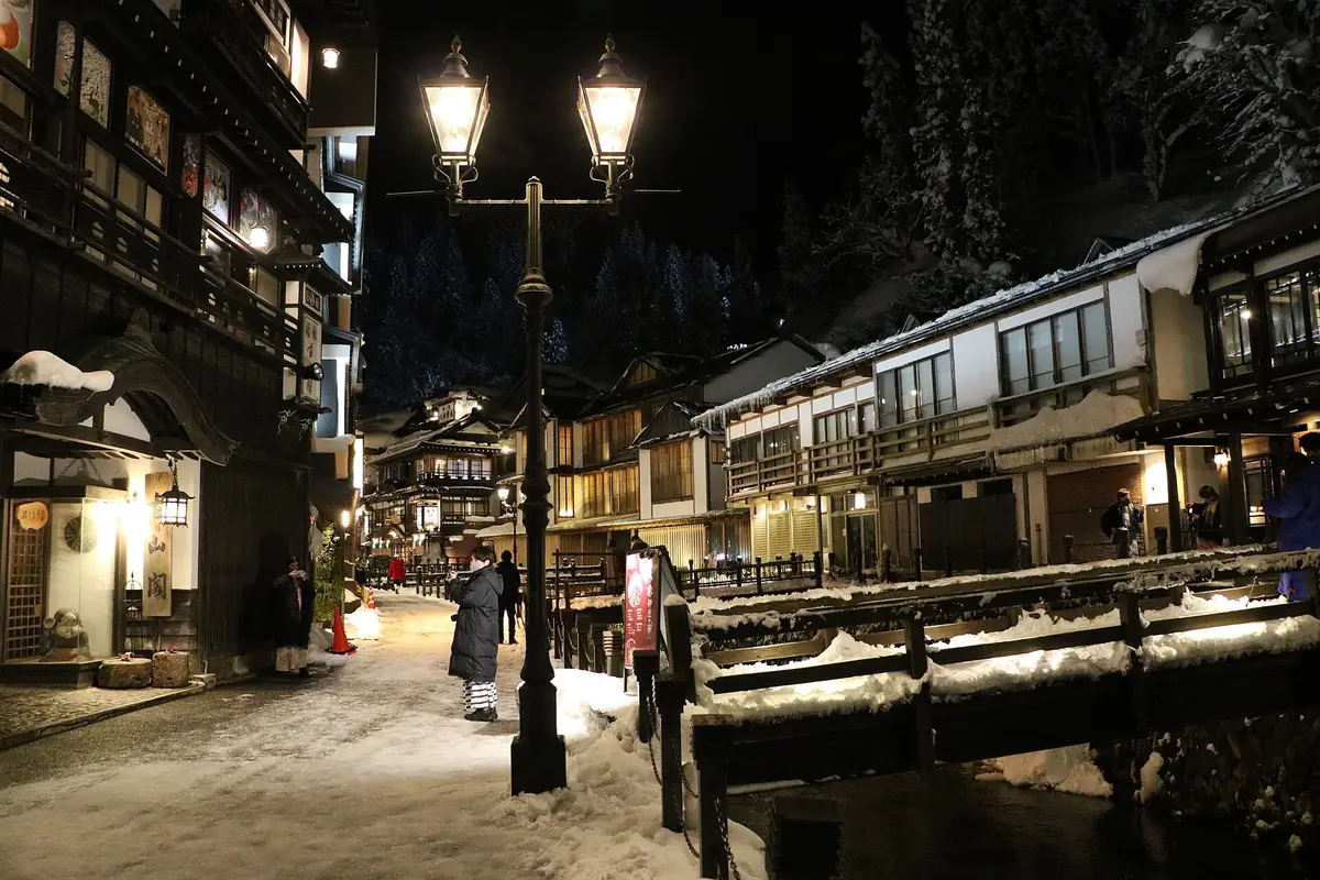 A twin gas lamp burning over the snow-packed street after dark in Ginzan Onsen, with the lit wooden ryokan lining the river behind it