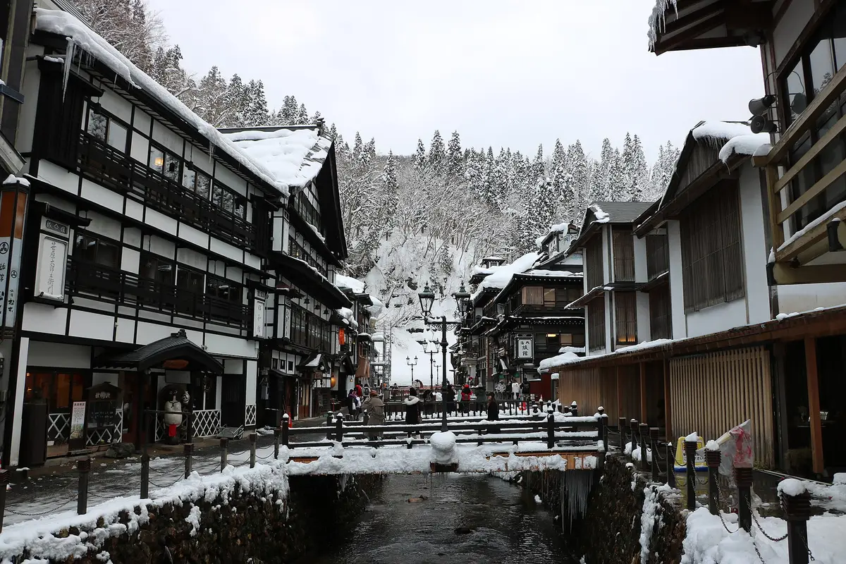 Looking up the valley on an overcast winter afternoon: the Ginzan River between two rows of snow-covered wooden ryokan, with visitors on the small bridge