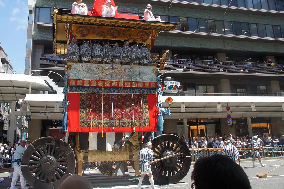 A tall Gion Matsuri float on a Kyoto street with musicians seated under its roof, two large wooden wheels below, and spectators lining the pavement