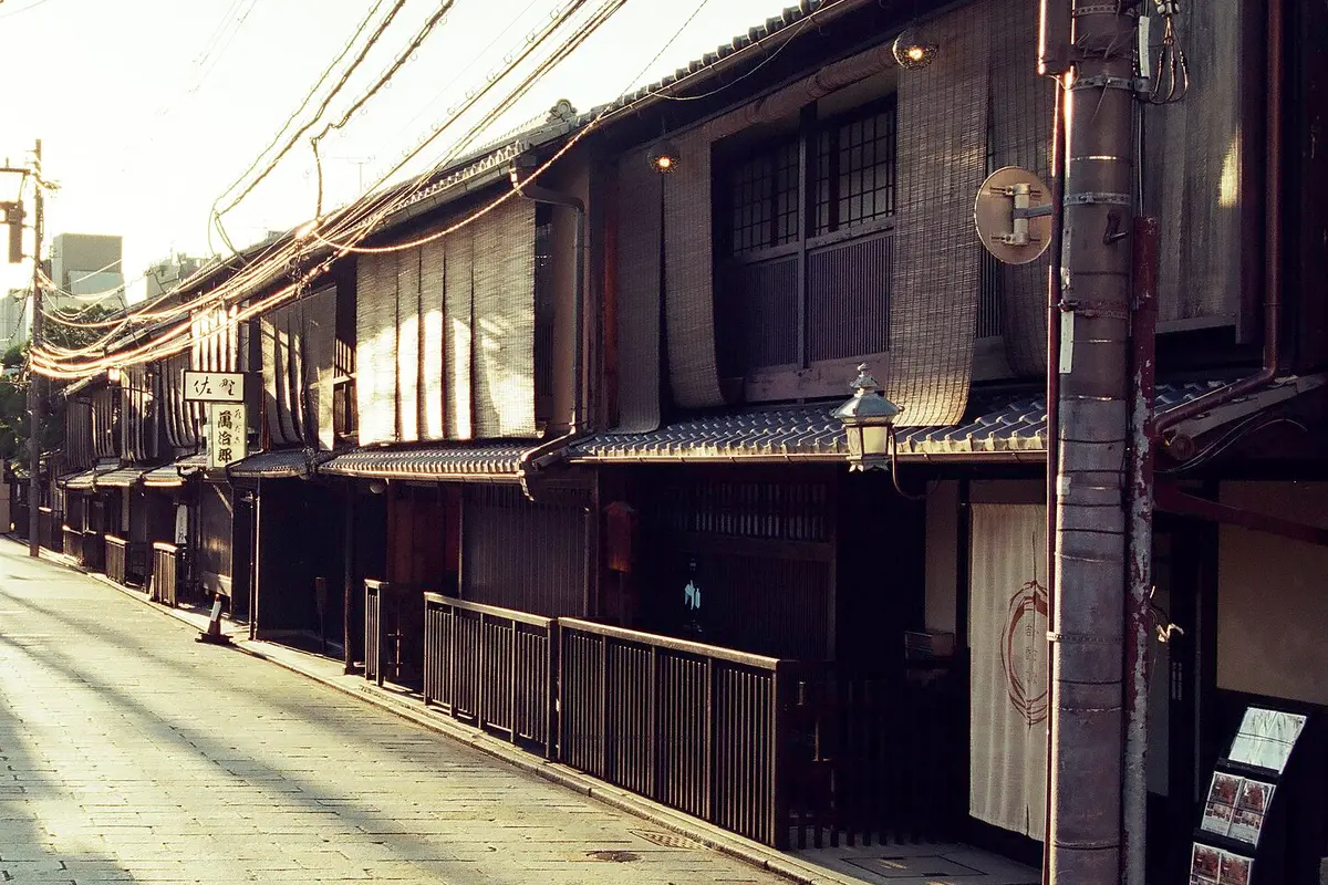 Một dãy nhà gỗ hai tầng ở Gion, Kyoto, với mành tre, một chiếc đèn lồng treo và con đường lát đá vắng người dưới ánh nắng thấp ấm áp.