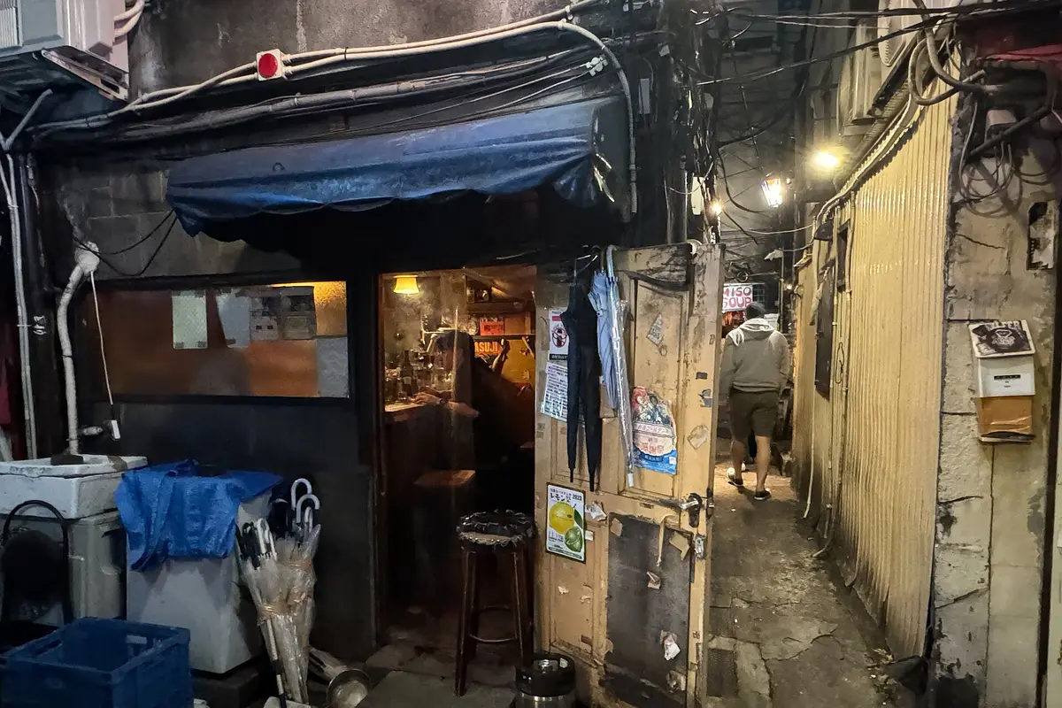 The open door of a tiny Golden Gai bar at night, with a wooden counter, a stool and shelves of bottles inside, and a narrow lane alongside