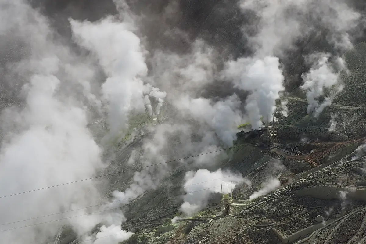 White steam rising from many vents across the bare volcanic slope of Ōwakudani, with a ropeway cable strung above the valley and stone terracing on the right
