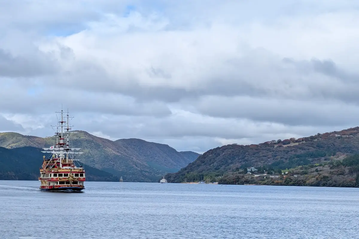 From the shore of Lake Ashi: a Hakone sightseeing pirate ship crossing the grey water under a heavily clouded sky, with wooded hills on both sides