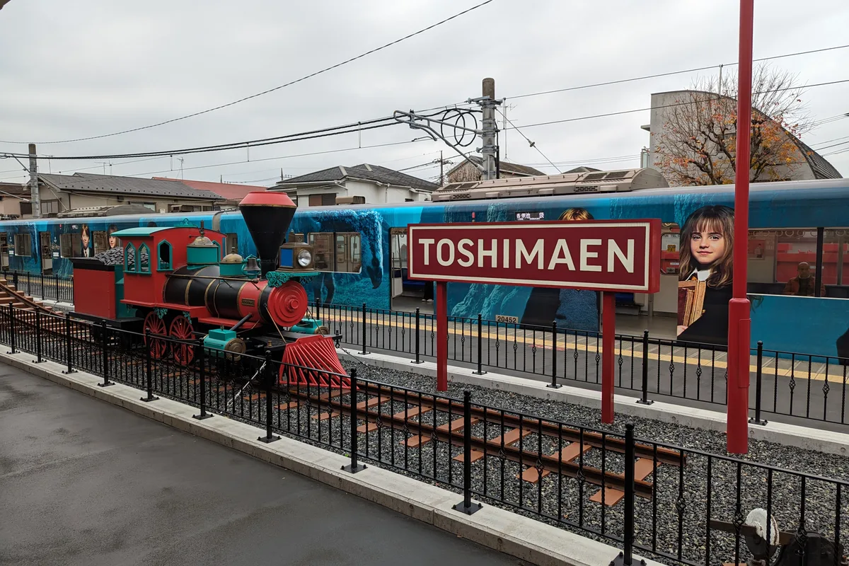 A miniature red steam engine sits beside the Toshimaen train platform, where a real commuter train wrapped in Harry Potter film artwork waits at the sign reading TOSHIMAEN
