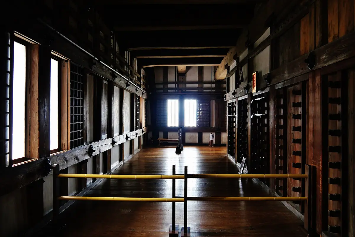 Ein leeres Stockwerk im Hauptturm der Burg Himeji mit blanken Holzdielen, Holzpfosten, Gitterfenstern, Waffenständern an der Wand und einem niedrigen Absperrgeländer im Vordergrund