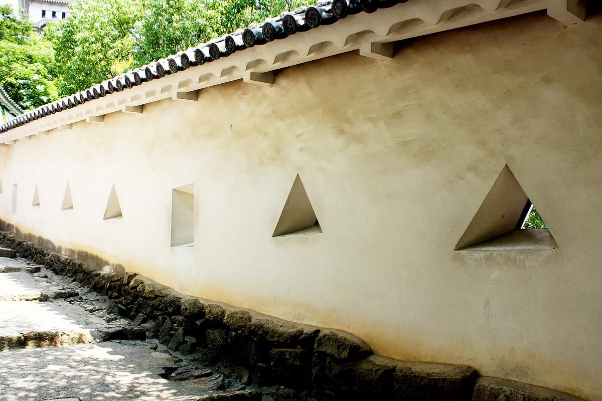 Weiße Putzmauer der Burg Himeji mit dreieckigen und quadratischen Öffnungen, oben ein Ziegeldach, unten ein steinerner Sockel und daneben ein Weg