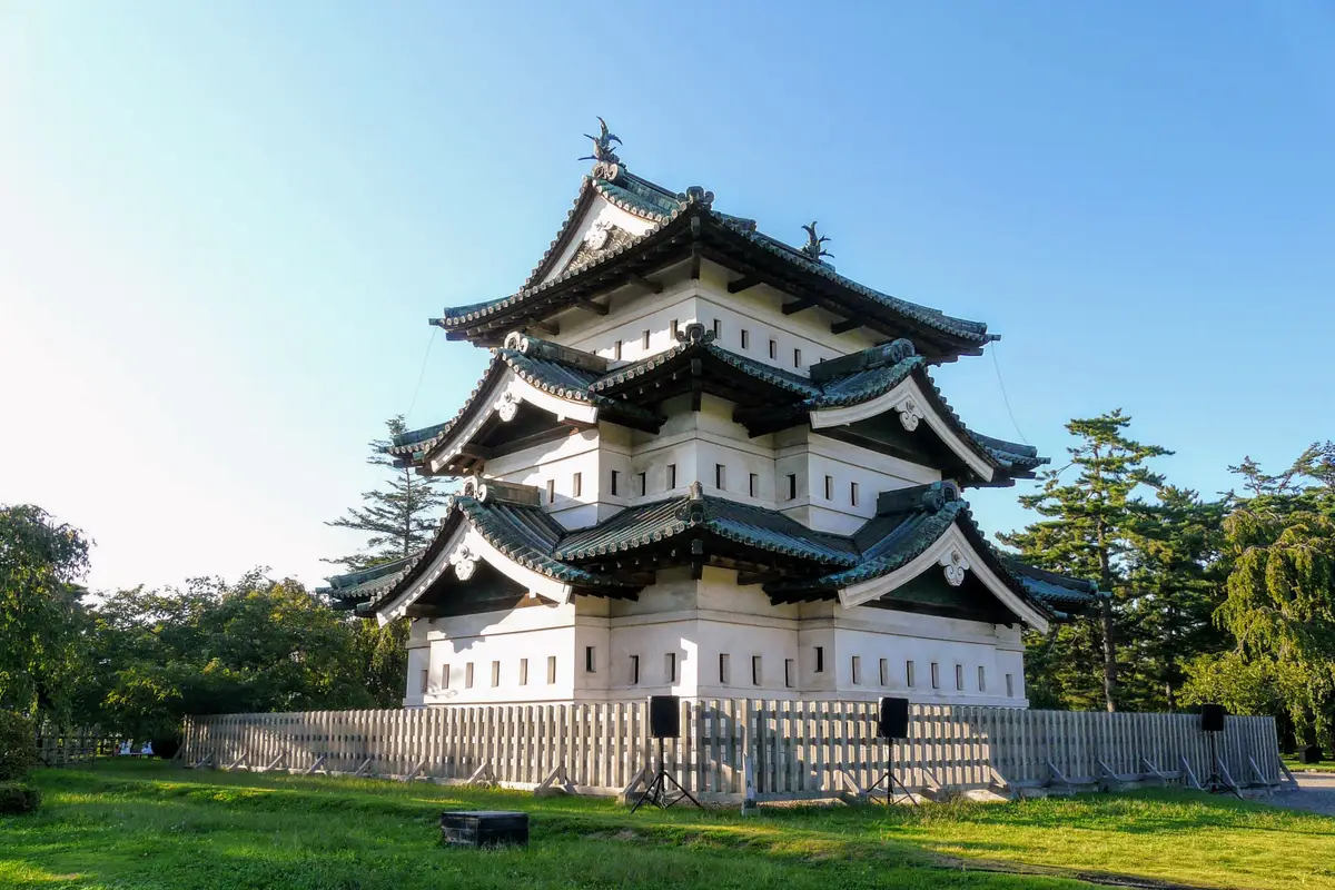 Über den Rasen gesehen: der kleine dreistöckige hölzerne Bergfried der Burg Hirosaki auf einer niedrigen provisorischen Plattform hinter einem Holzzaun