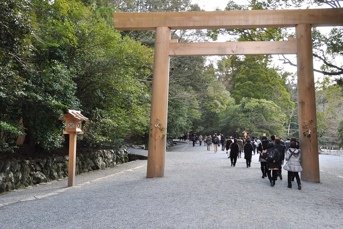 Visitors walking away across the gravel approach beneath a plain wooden torii, with forest on both sides, at Ise Jingu