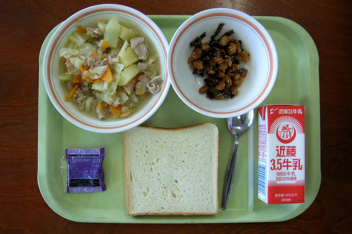 从正上方拍摄的日本小学营养午餐餐盘，上面有肉类蔬菜汤、一小碗调味豆子和海藻、一片吐司、一包果酱、一支汤匙和一盒牛奶
