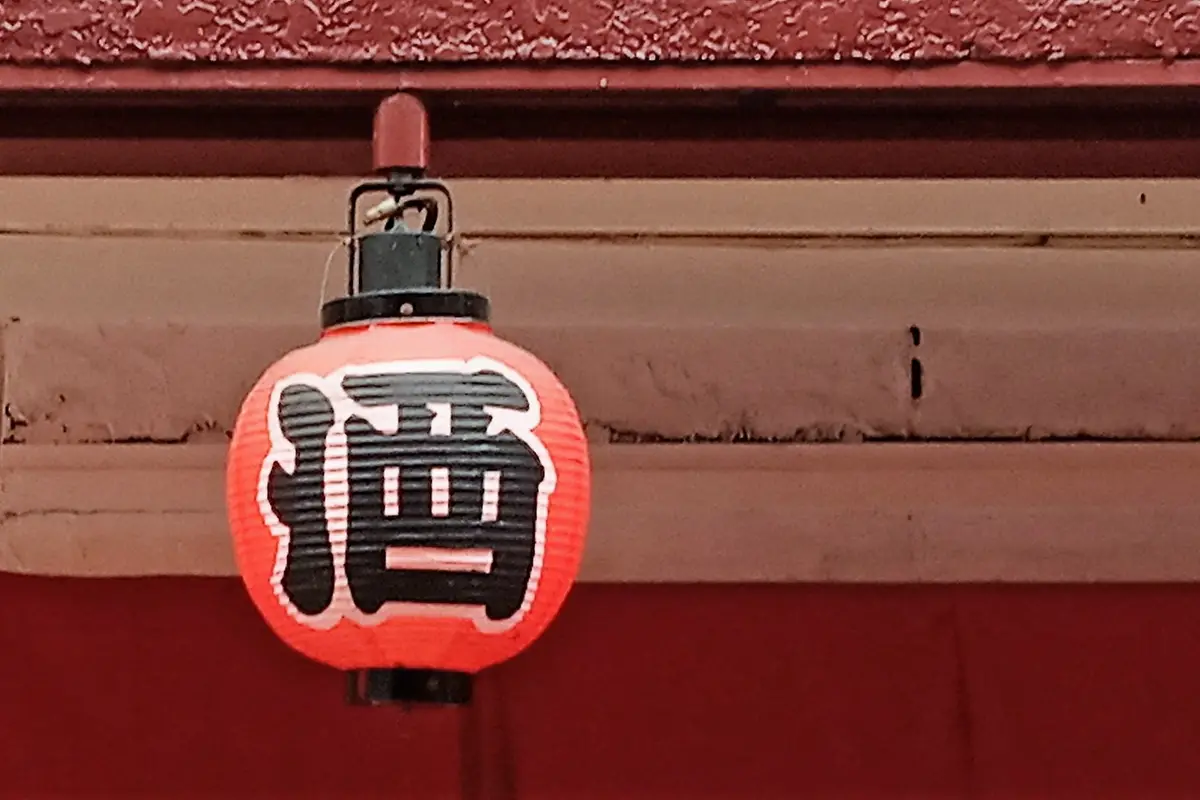 A round red paper lantern painted with the character for sake, hanging under the eaves of an izakaya above a deep red cloth curtain