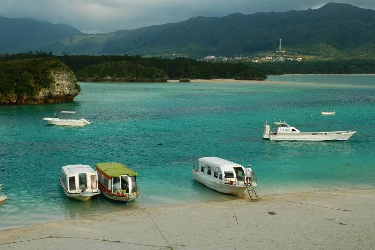 Glass-bottom tour boats moored at the white sand beach of Kabira Bay, with turquoise water and wooded hills beyond