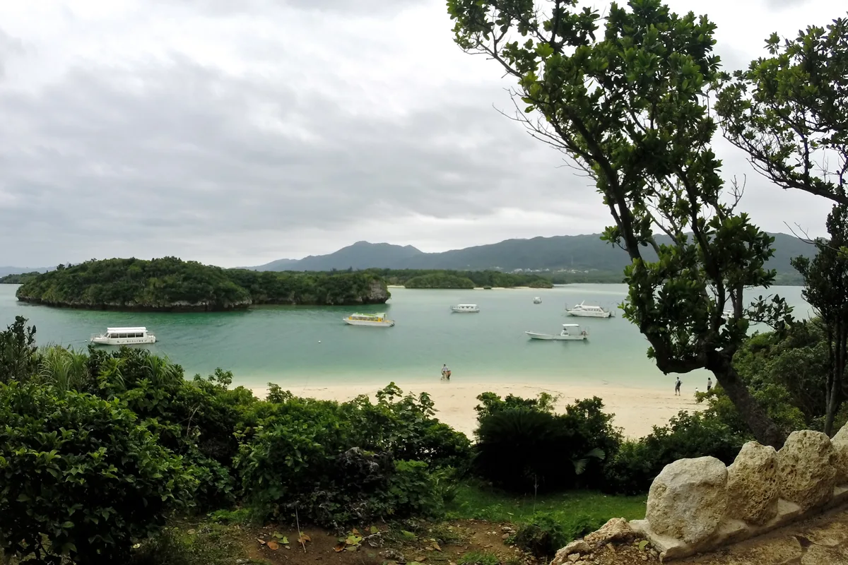A wide view of Kabira Bay under cloudy skies, with several tour boats anchored offshore and visitors standing on the sandbar