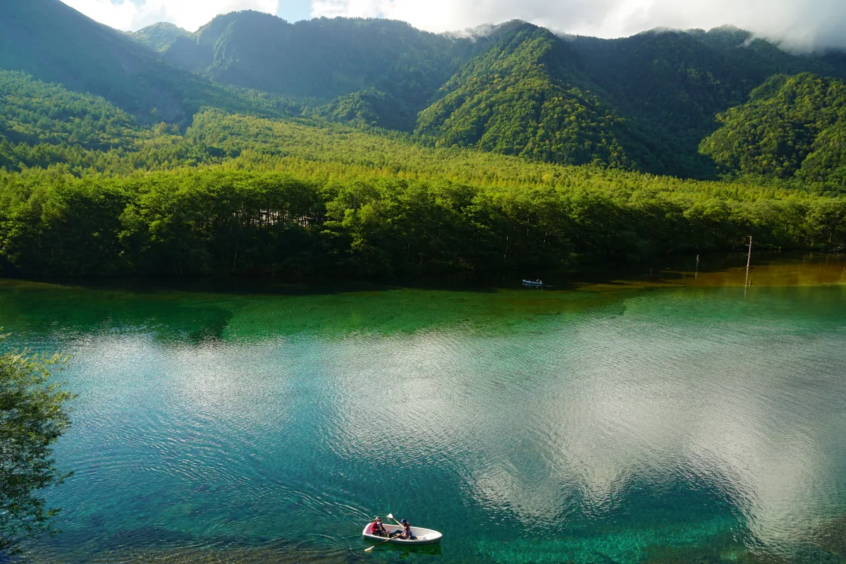 A small rowboat crossing the glass-clear green water of Taisho Pond in Kamikochi, forested mountains rising behind