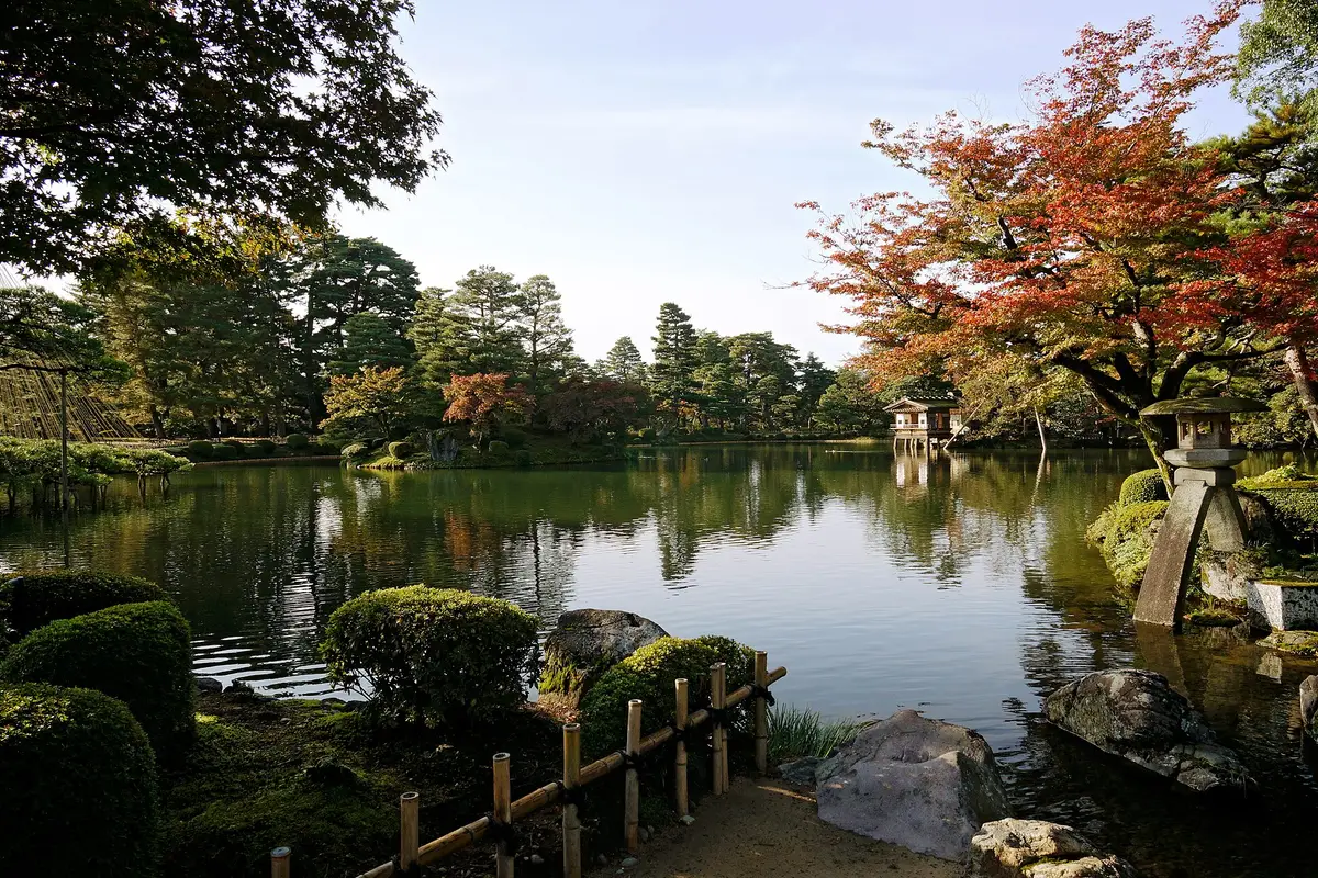Across Kasumigaike pond at Kenroku-en: clipped pines, mossy banks, an autumn maple and a small wooden pavilion standing over the water on the far shore