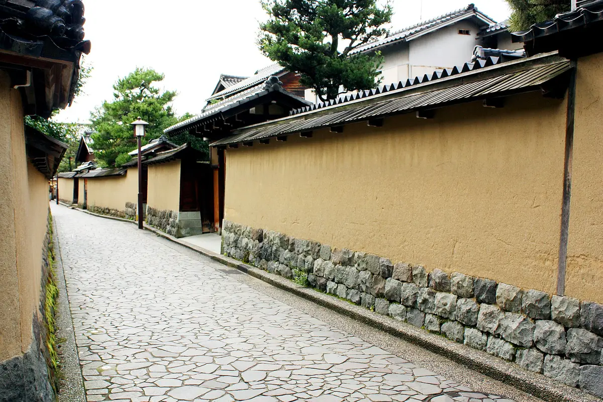 Ochre earthen walls topped with wooden eaves line a stone-paved lane in the Nagamachi samurai district of Kanazawa, with no one on the paving