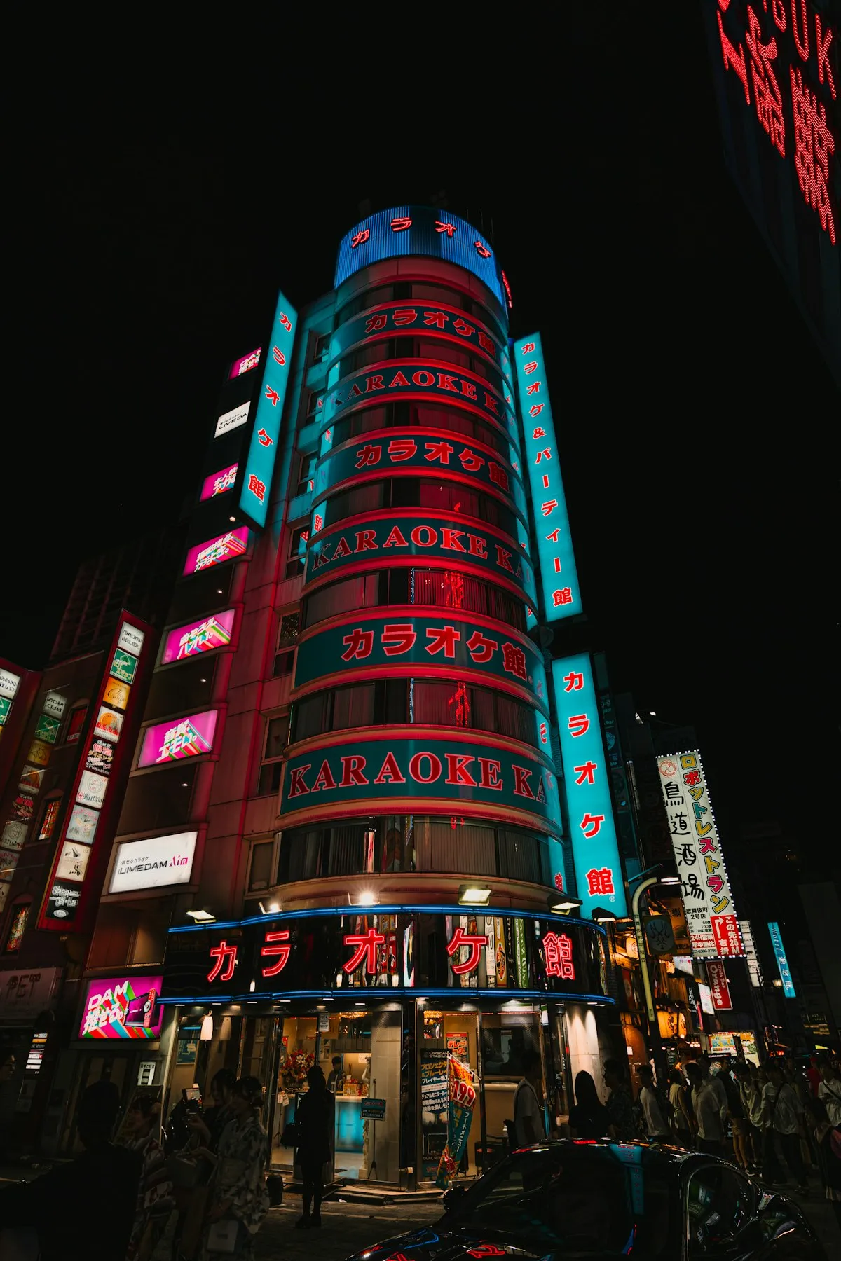 A multi-story karaoke building in Tokyo glowing with neon signs in Japanese and English at night