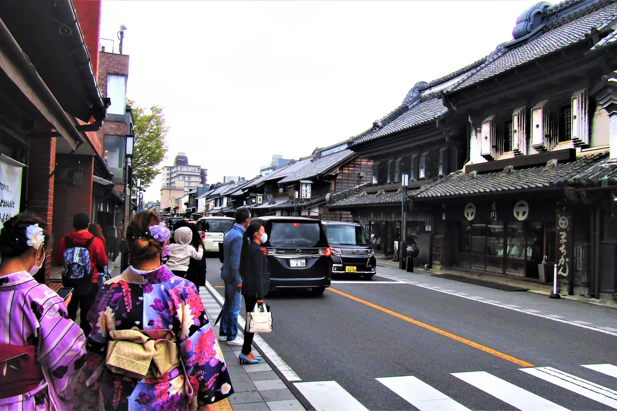 A narrow street in Kawagoe's Kurazukuri district lined with dark warehouse-style buildings, with cars and pedestrians sharing the same road