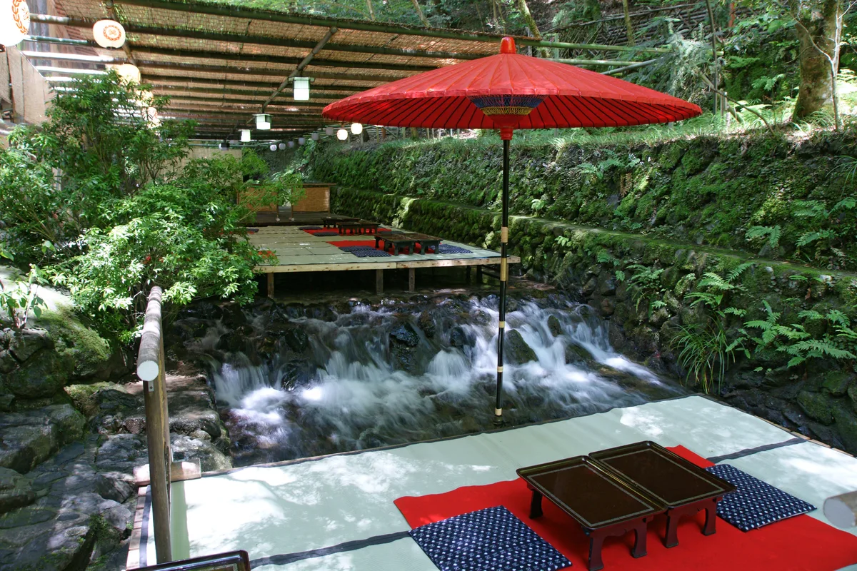 A low dining platform on stilts set out over the rushing Kibune stream, shaded by a red paper parasol and a reed roof, surrounded by mossy green banks