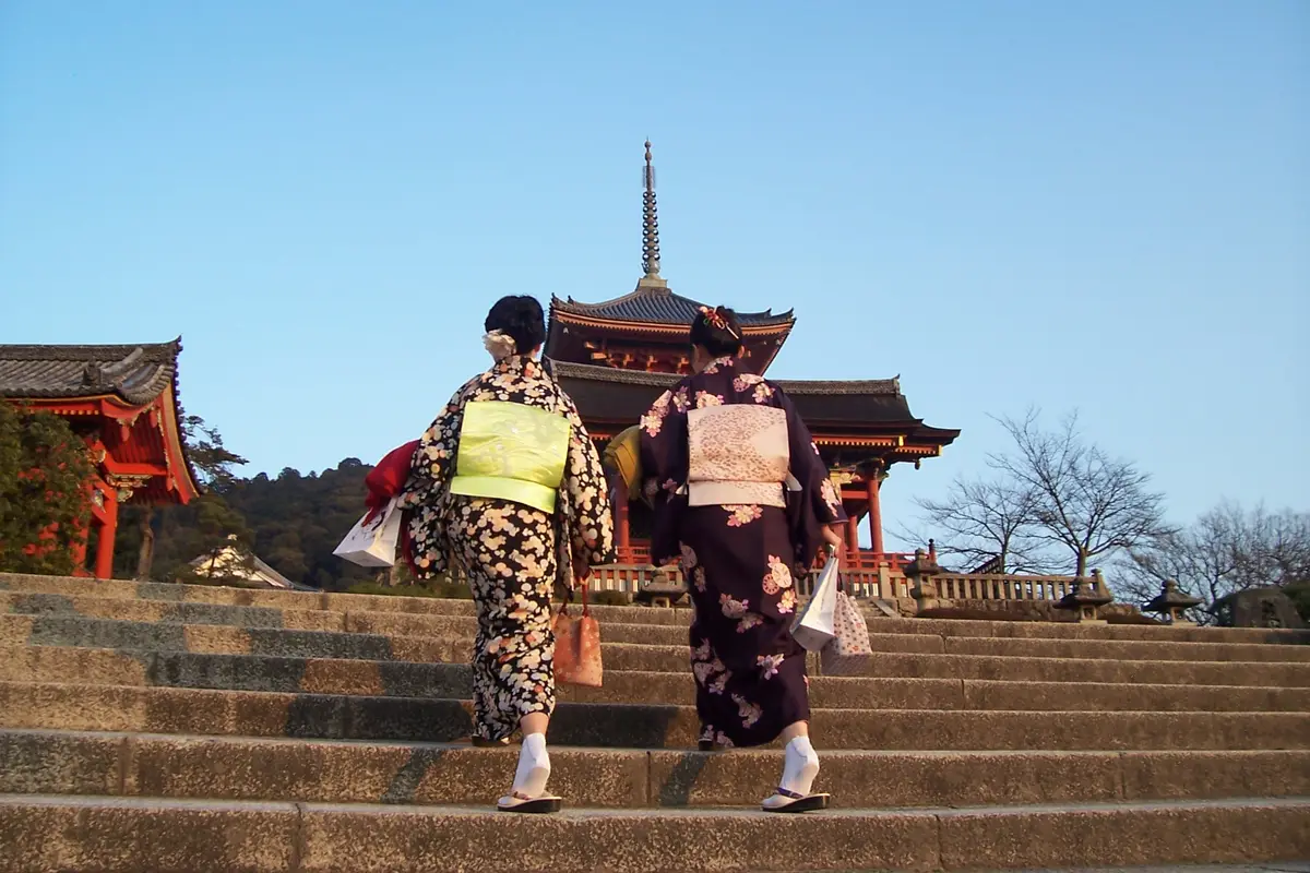 Seen from behind, two people in kimono climbing the stone steps toward the vermilion gate and three-storied pagoda of Kiyomizu-dera in Kyoto