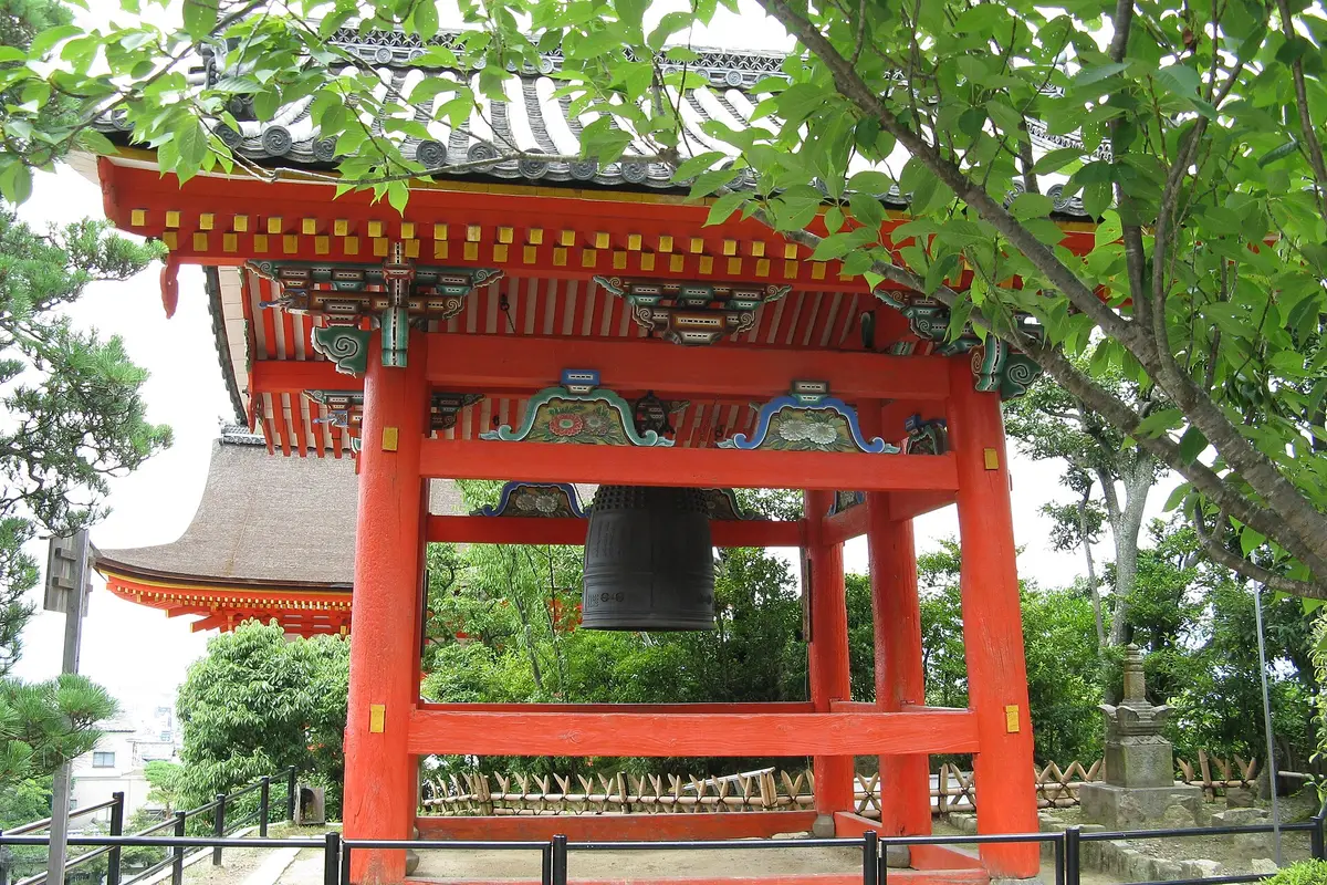 Der zinnoberrote Glockenturm von Kiyomizu-dera zwischen grünem Laub, die große Bronzeglocke hängt unter bemalten Konsolen