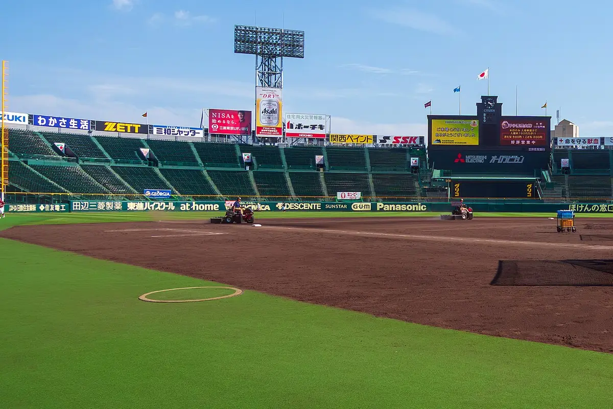 The bare brown infield soil of Hanshin Koshien Stadium seen from field level, with the green outfield in front and the empty stands behind