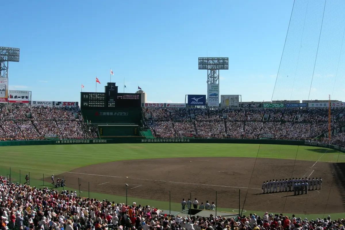 Hanshin Koshien Stadium packed to the upper deck under a blue sky, with a row of players lined up along the foul line during the national high school final