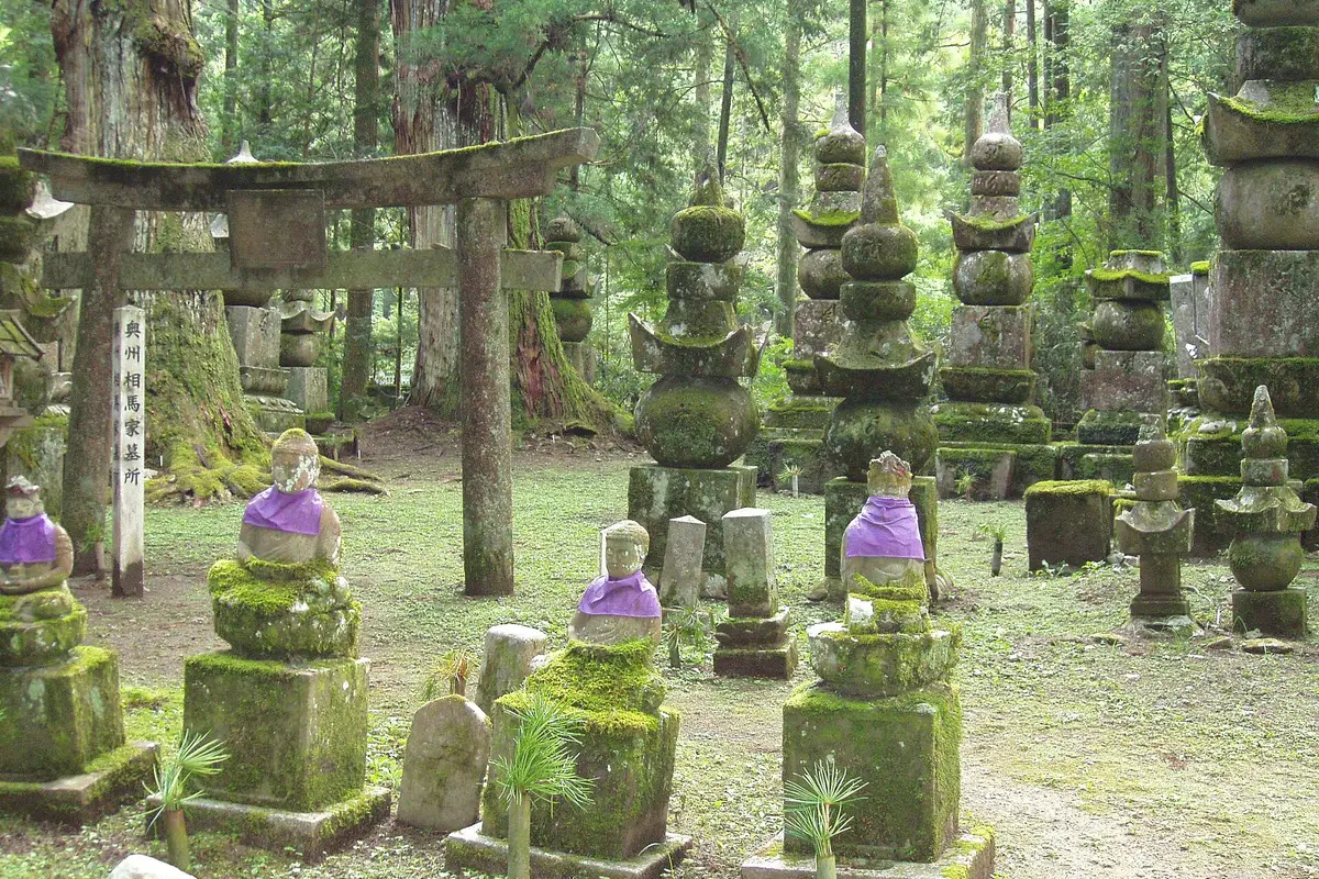 Moss-covered memorial stones, small stone statues in cloth bibs and a weathered stone torii standing among tall cedars in the Okunoin cemetery on Koyasan