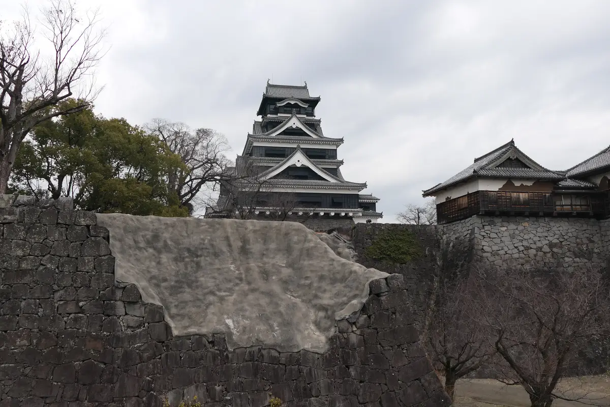 Kumamoto Castle's reconstructed main keep rising behind a stone wall whose collapsed face has been sealed in grey while it waits for repair