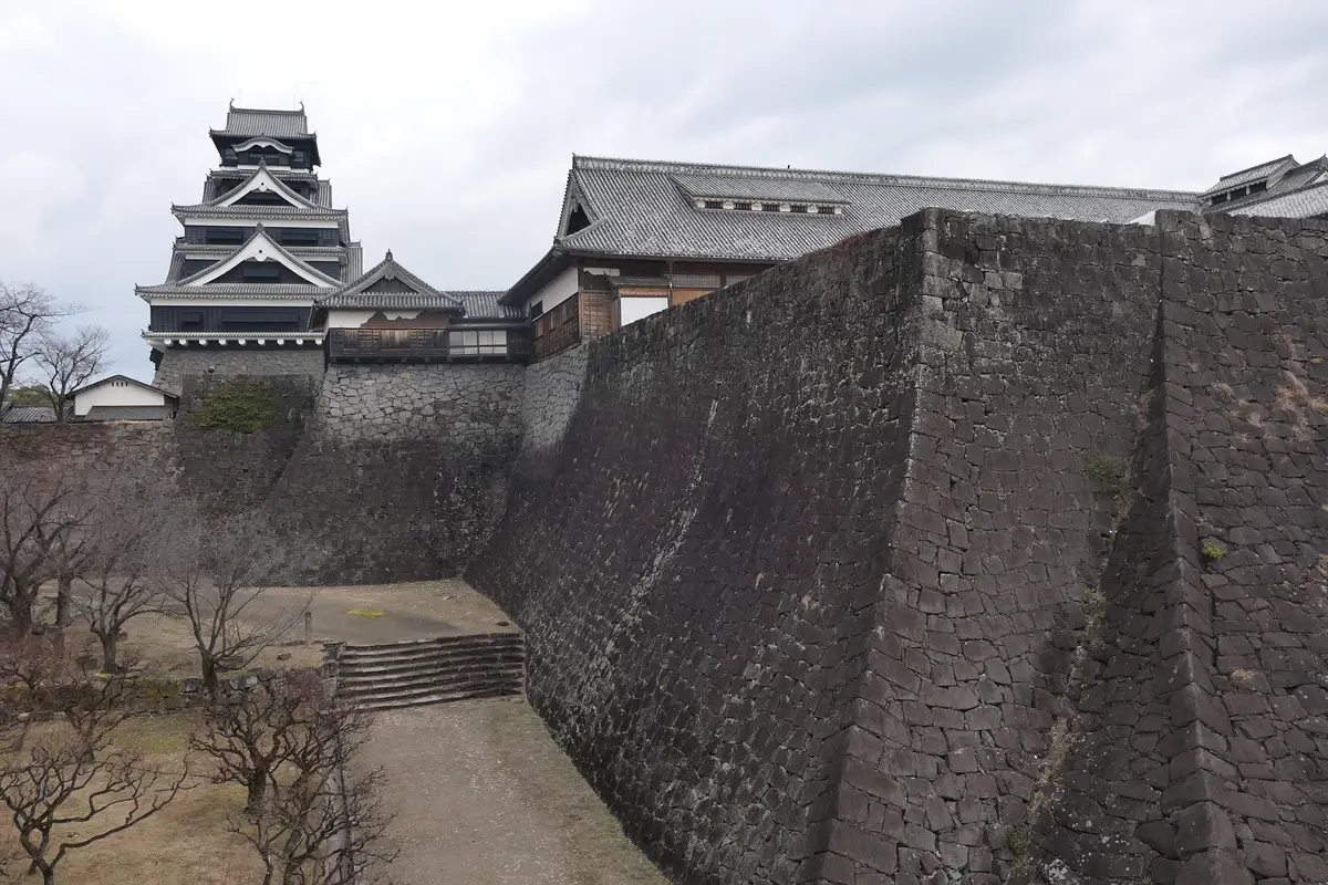 A long dark stone rampart of Kumamoto Castle curving upward and running toward the main keep, seen from the grounds at its foot