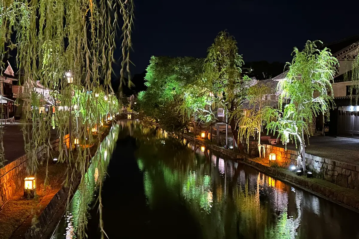 Visto da un ponte dopo il tramonto: i salici illuminati e la luce delle lanterne si riflettono nell'acqua ferma del fiume Kurashiki, con il camminamento lungo le rive di pietra deserto
