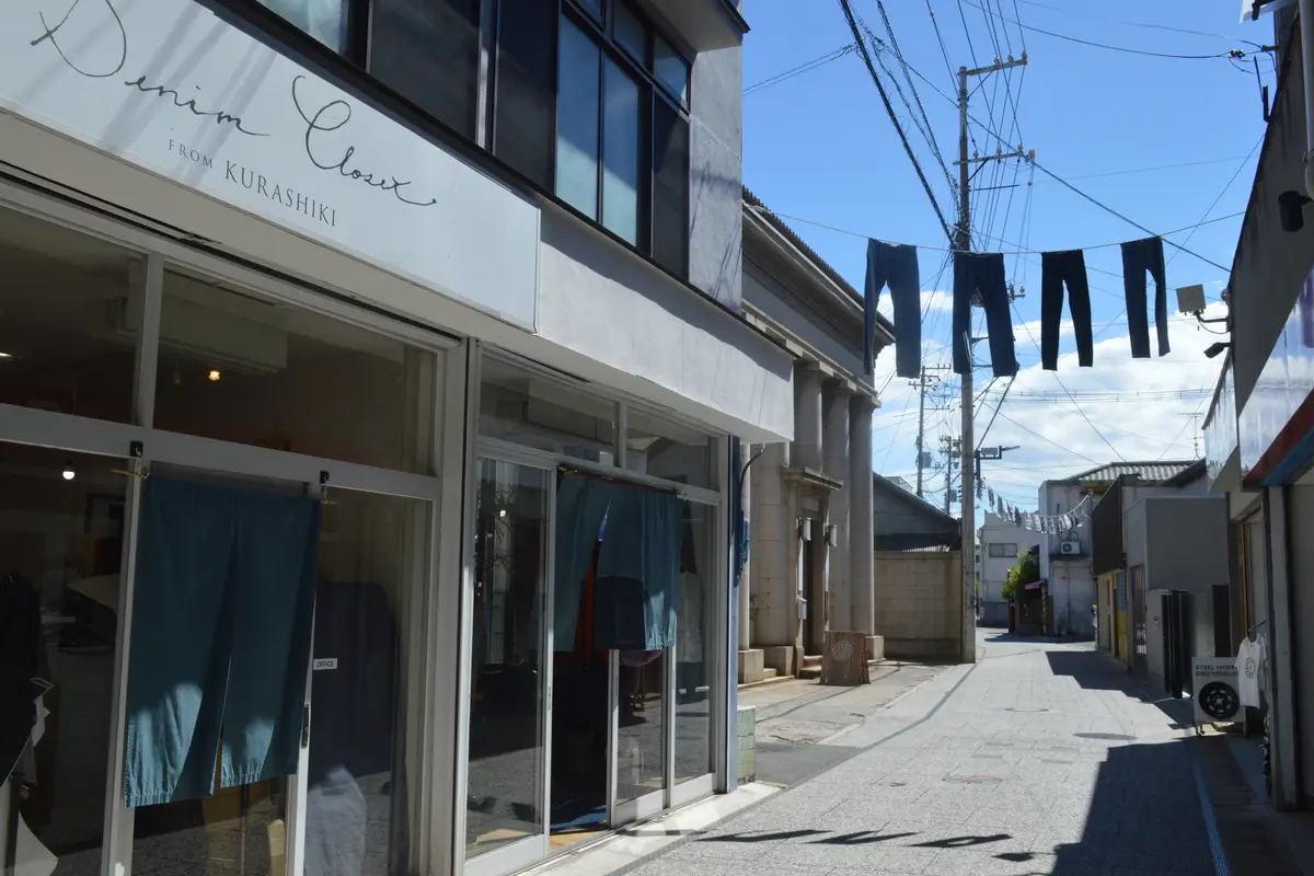 La Jeans Street nel quartiere di Kojima, a Kurashiki: denim indaco appeso in alto sopra una strada vuota, accanto a un negozio con l'insegna Denim Closet from Kurashiki