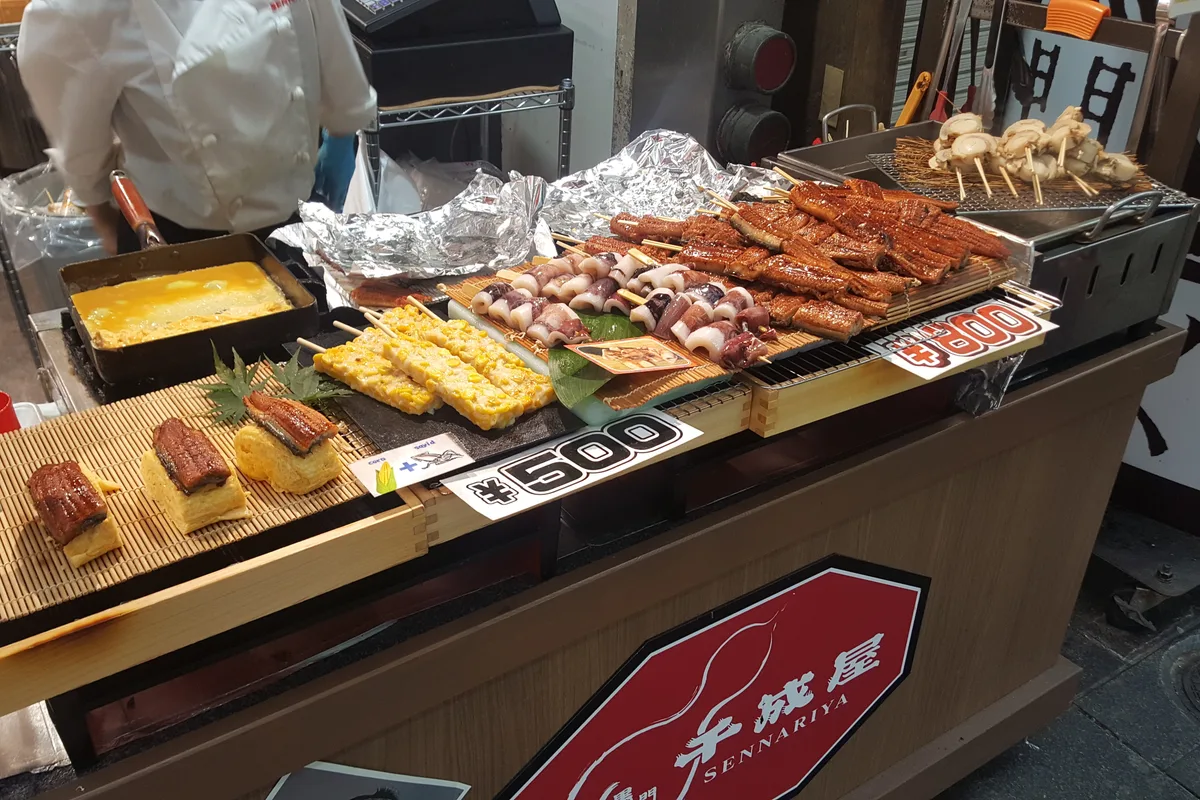 แผงอาหารทะเลที่ตลาดคุโรมง มีปลาไหลย่าง ไม้ปิ้ง และทามาโกยากิวางเรียงอยู่ข้างป้ายราคาแบบพิมพ์ที่เขียนว่า ¥500 และ ¥800