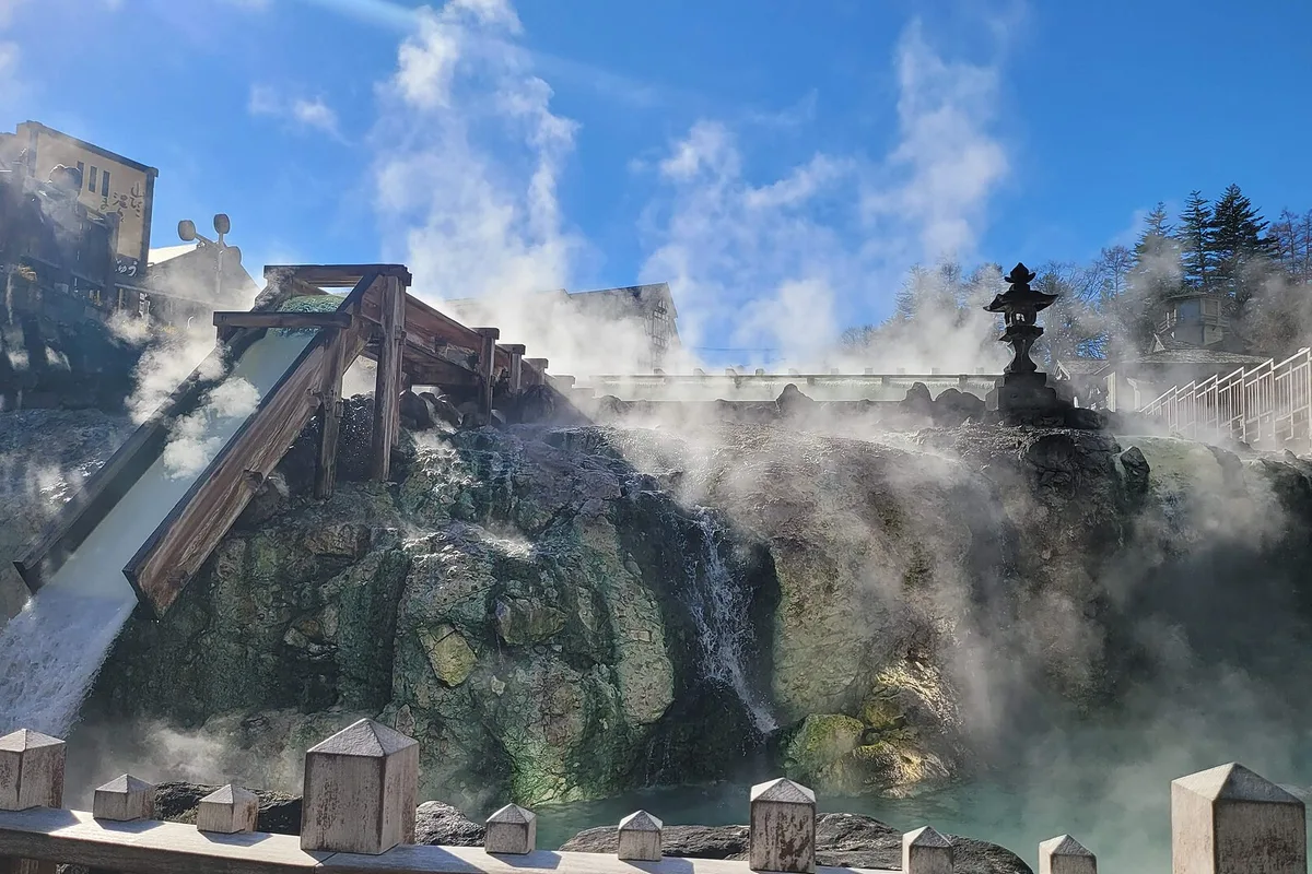 Close view of the wooden cooling channel at Yubatake pouring into mineral-stained rock, steam rising off the greenish acidic water