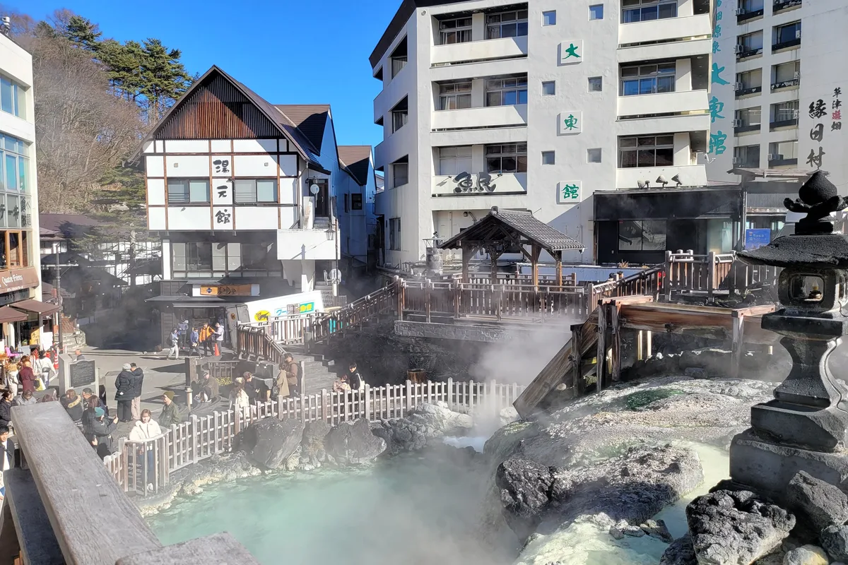 View down over Yubatake with its wooden channels and stone lantern, ryokan and shop buildings crowding the slope, and visitors gathered along the railings