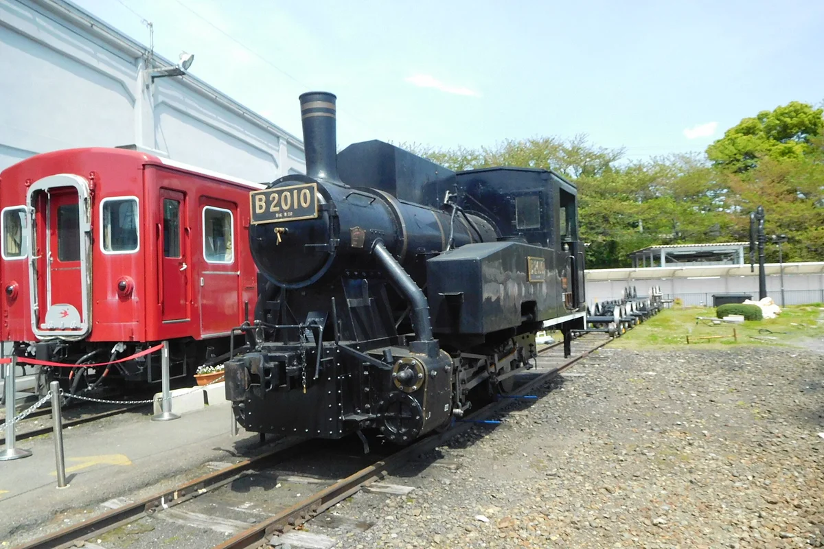 Black steam locomotive B2010 beside a retired red train car at the Kyoto Railway Museum roundhouse