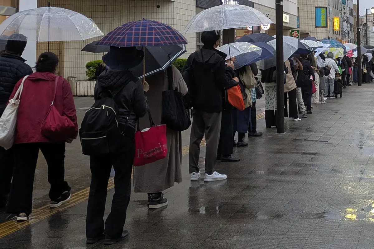 A line of people holding umbrellas, seen from behind, waiting at a bus stop outside a station in Japan on a rainy evening