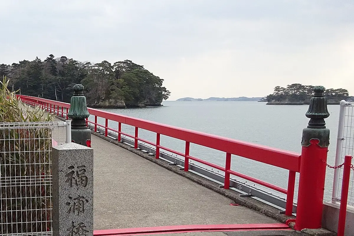 La ringhiera vermiglia del Ponte Fukuura si allunga sull'acqua verso Fukuurajima, l'isola coperta di pini, con un pilastro di pietra all'ingresso
