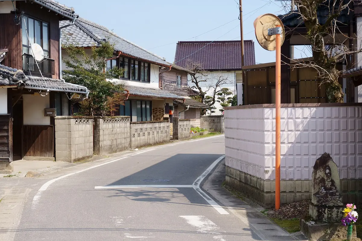 A quiet residential street in a small Japanese town, with tiled roofs, low block walls and a small roadside stone figure with fresh flowers