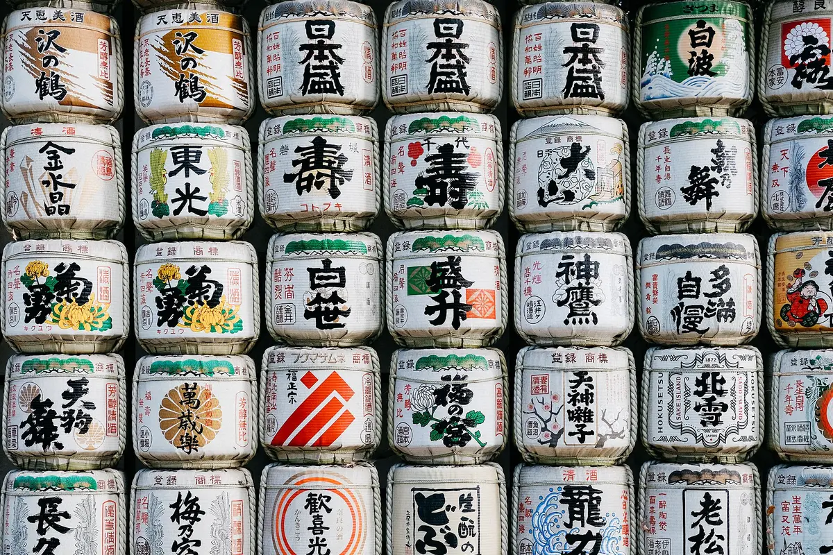 A wall of straw-wrapped sake barrels stacked in rows, each painted with a brewery's name in bold characters