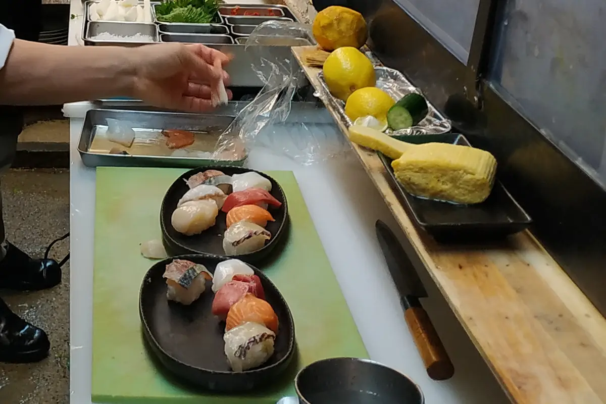 A sushi chef's forearm and hand working at the counter, with two plates of nigiri on a cutting board beside lemons and a knife