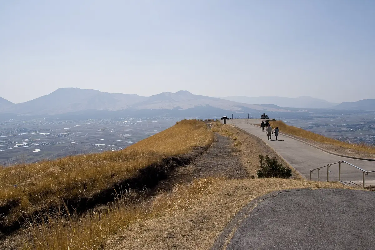 Visitors walking the northern rim path at Daikanbo, with the flat floor of the Aso caldera and its fields and towns spread far below