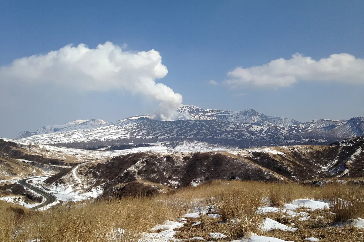 A white plume of volcanic gas rising from the summit of Nakadake, seen from a distance across the snow-dusted caldera
