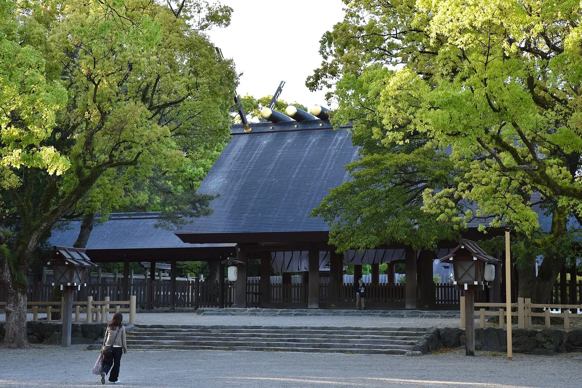 A visitor approaches the wooden worship hall of Atsuta Jingu in Nagoya, framed by a dense grove of tall green trees