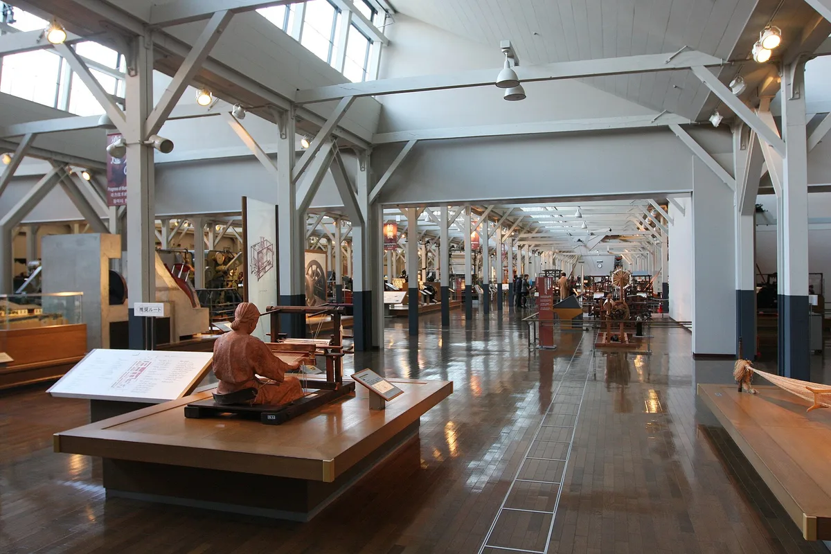 A seated mannequin works a hand loom in the foreground, with rows of vintage power looms and textile machinery receding under the wood-framed roof of the Toyota Commemorative Museum's Textile Machinery Pavilion in Nagoya