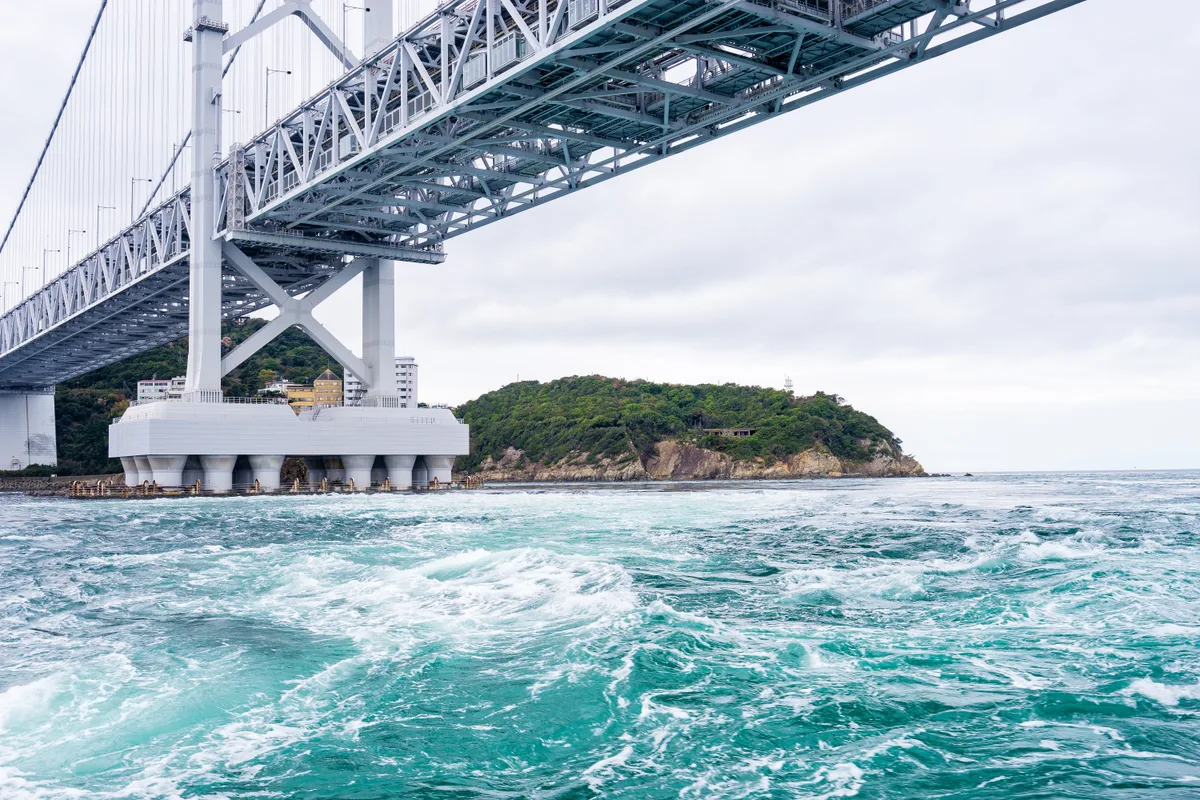 Il ponte Onaruto visto dall'acqua, con la corrente turchese vorticosa proprio sotto il pilone e un'isola verde sullo sfondo