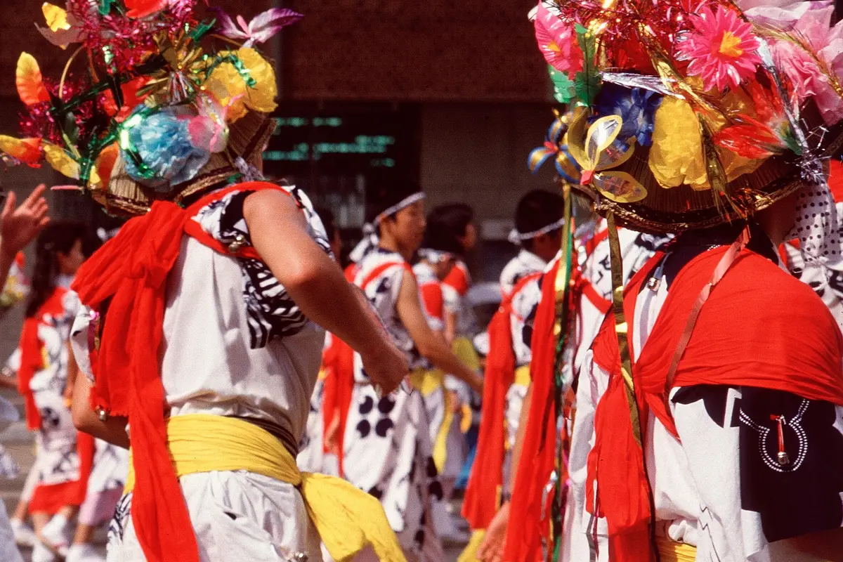 Two haneto seen from behind at the Aomori Nebuta Festival, in flower hats, red shoulder sashes and yellow waist sashes over white robes, with small bells at the shoulder